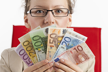 Frau mit Brille blickt über Fächer aus Banknoten