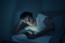 Addicted man chatting and surfing on the Internet with smart phone late at night in bed. Bored, sleepless and tired in dark room with moody light. In insomnia and mobile addiction concept.