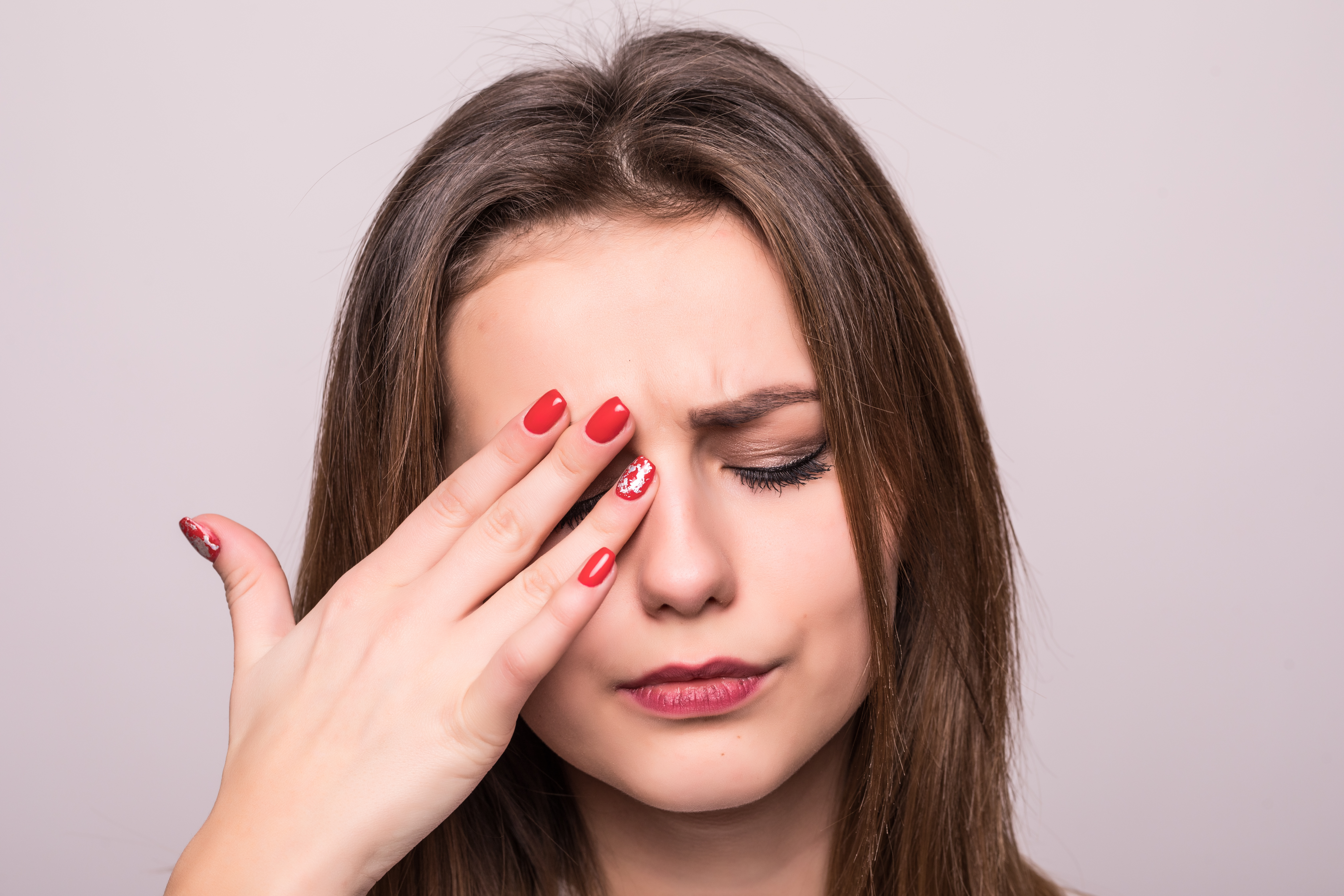 Pain. Tired Exhausted Stressed Woman Suffering From Strong Eye Pain isolated on gray. Portrait Of Beautiful Young Female Feeling Sick, Having Headache, Nose Pain And Touching Painful Eyes.